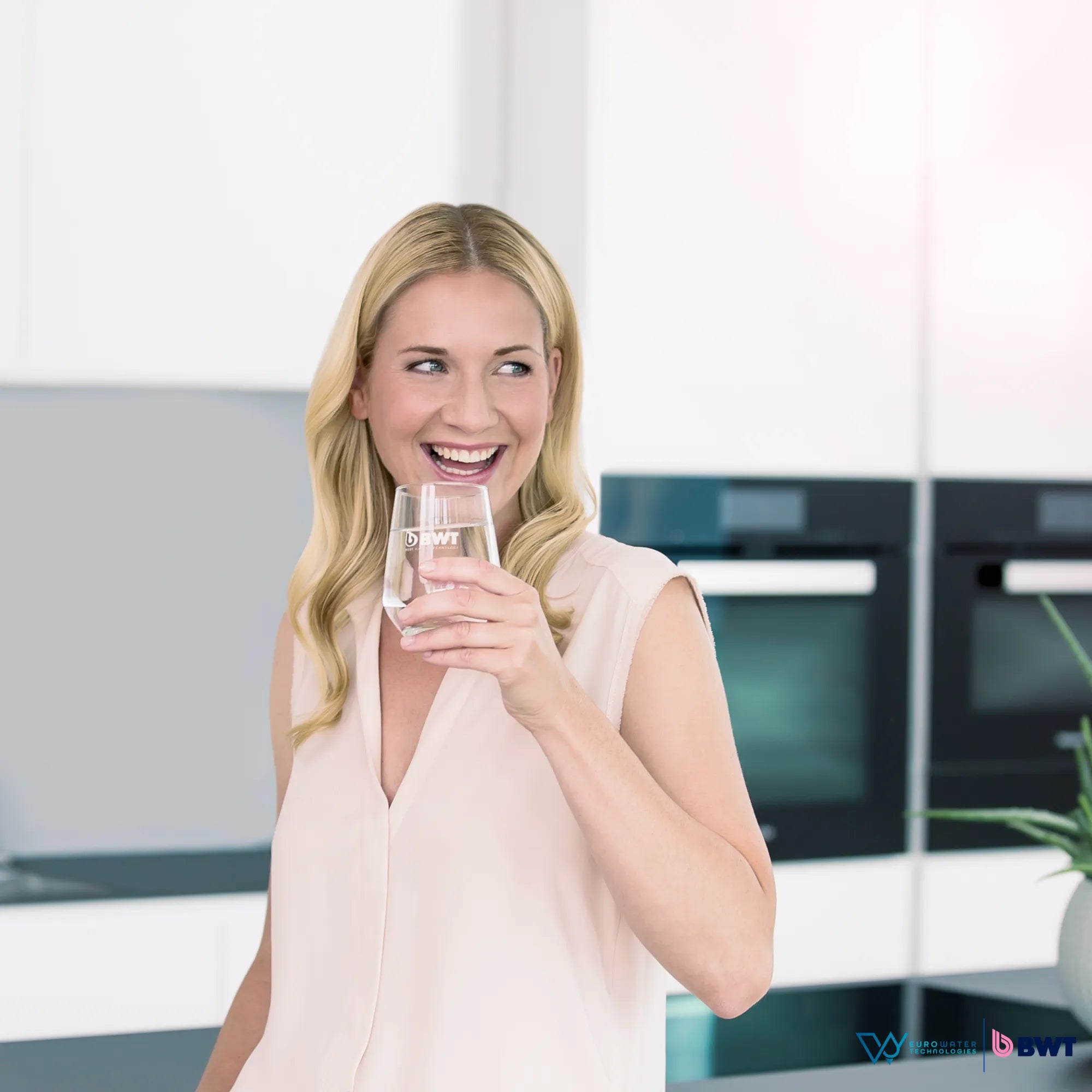 Mujer rubia sonriente sosteniendo un vaso con agua, de fondo hay una cocina y refrigerador. Acompañada de los logos de Eurowater y BWT.